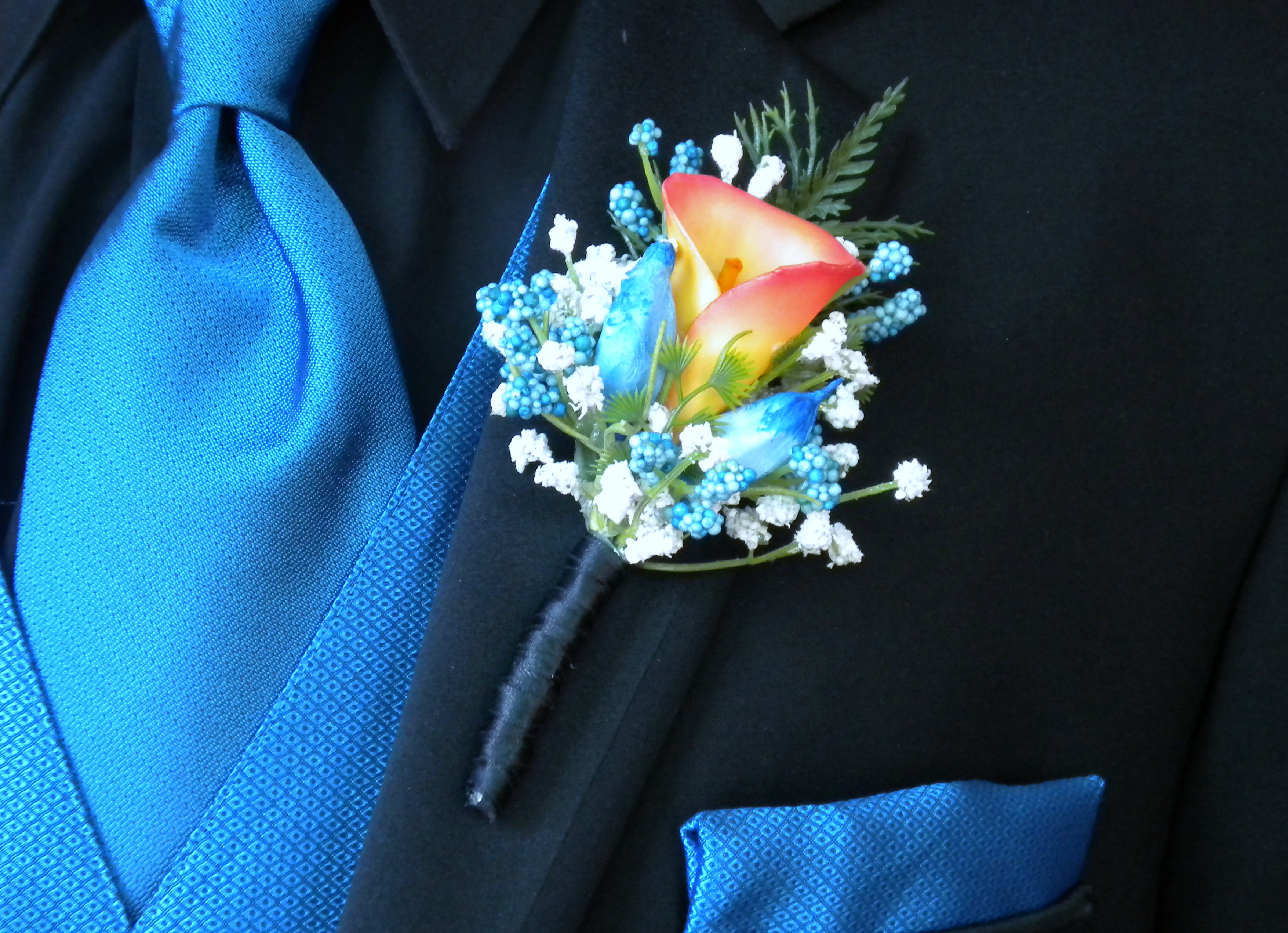 orange calla lily boutonniere with blue flowers and baby's breath against a black tuxedo and blue vest